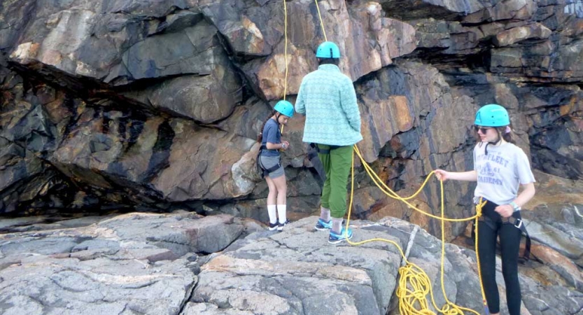 A group of students wearing helmets and other safety gear stand at the bottom of a rock facing, holding ropes. 